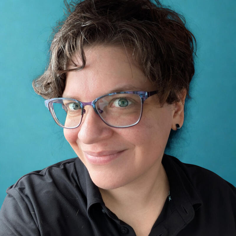 A picture of Natacha, the owner of Cosmic Spider. She has short brown hair and blue eyes. She is smiling, wearing a black polo shirt, and purple and blue glasses.